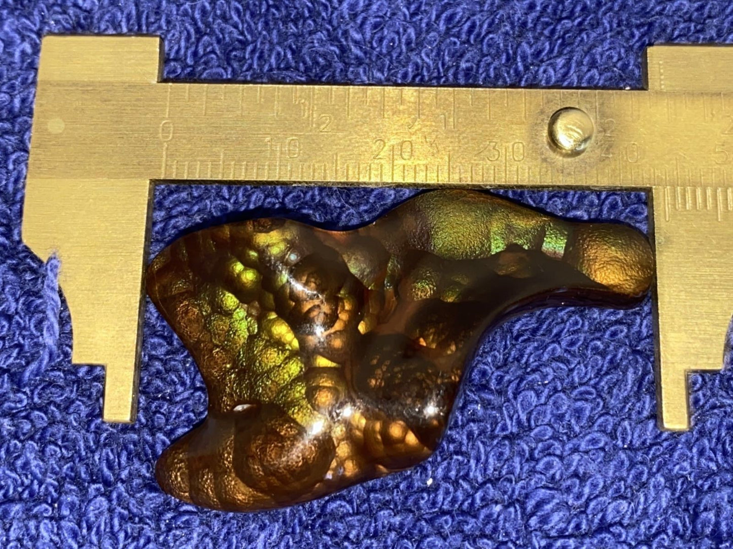 Multi-colored stone with green flecks on a blue textured background, measured by a brass ruler.