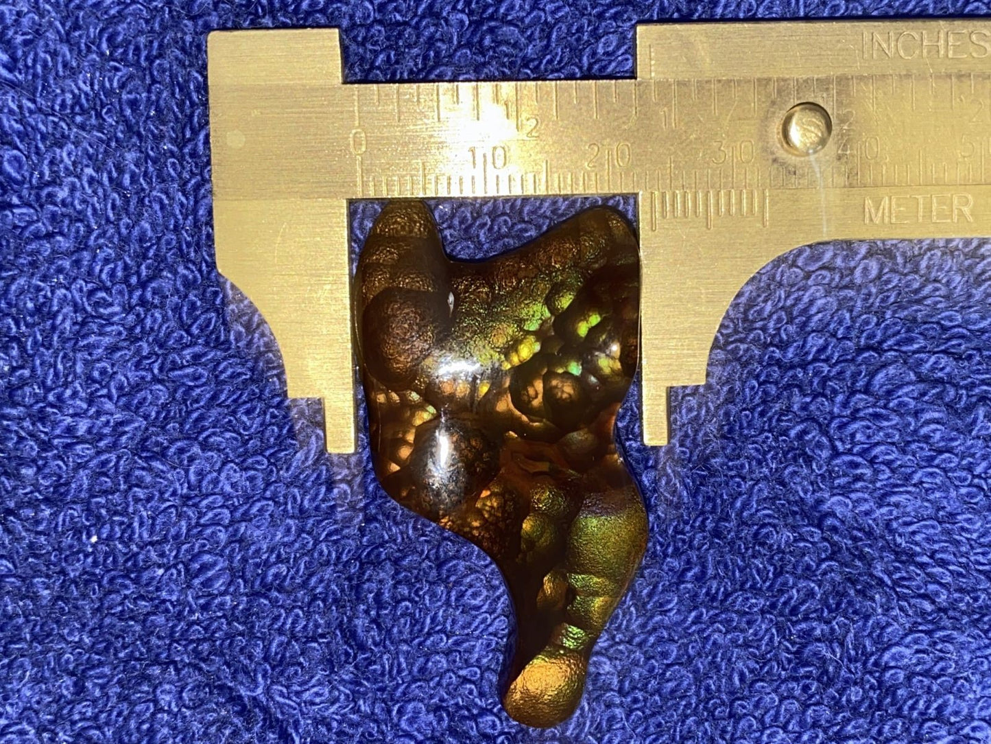 Multi-colored gem with green flecks on a blue textured background, measured by a gold ruler.