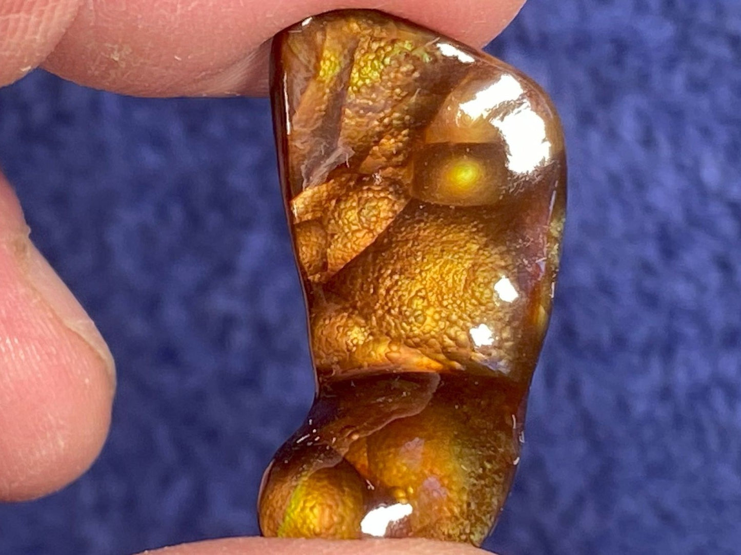 Close-up of a hand holding a polished Deer Creek Fire Agate Gemstone against a blue textured background