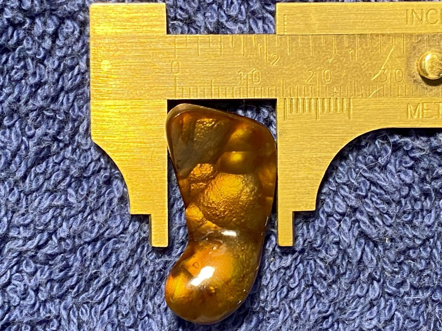 Deer Creek Fire Agate Gemstone being measured with a gold caliper on a textured blue background