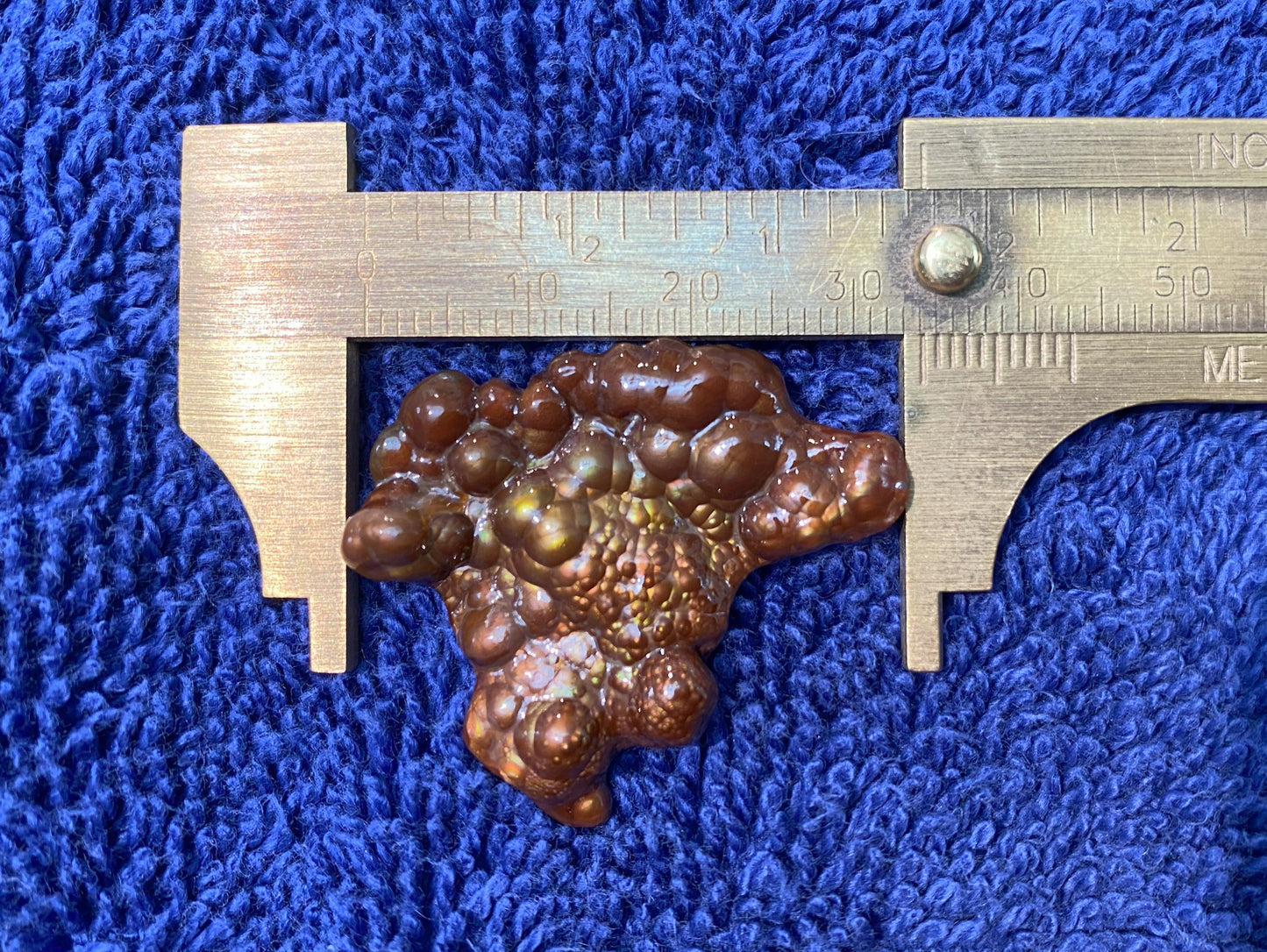 Deer Creek Fire Agate Gemstone Mineral Specimen being measured with a metallic caliper on a blue textured background