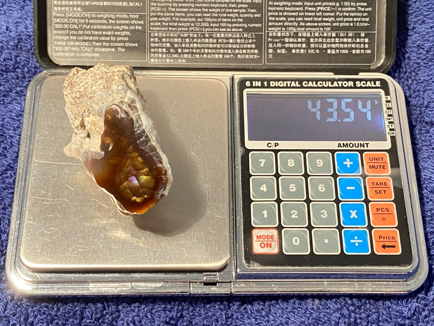Digital calculator scale with a large windowed piece of Aguascalientes Fire Agate rough on a blue surface
