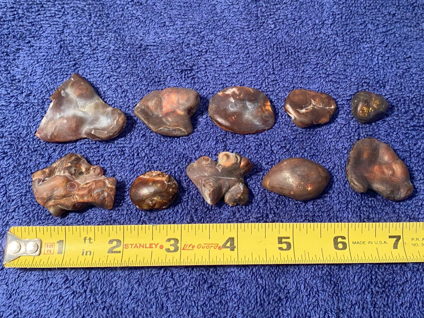 Collection of 10 fire agate gemstone preforms on a blue surface with a measuring tape for scale.