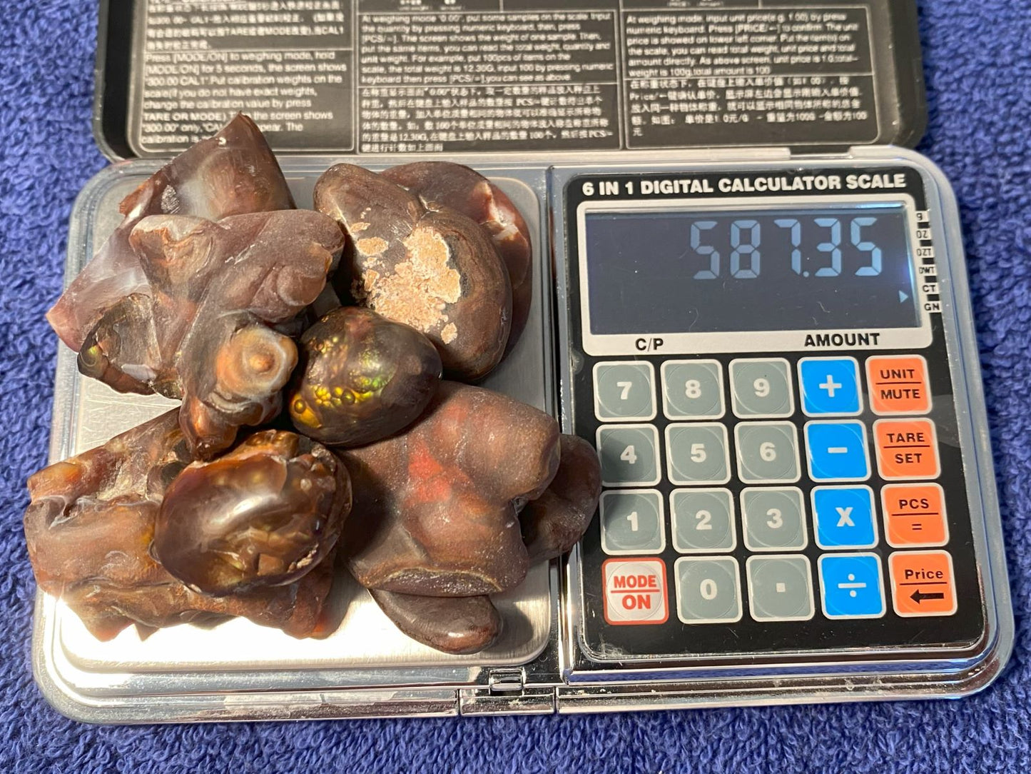 Digital calculator scale with fire agate preform gemstones displaying a weight of 587.35 carats on a blue surface