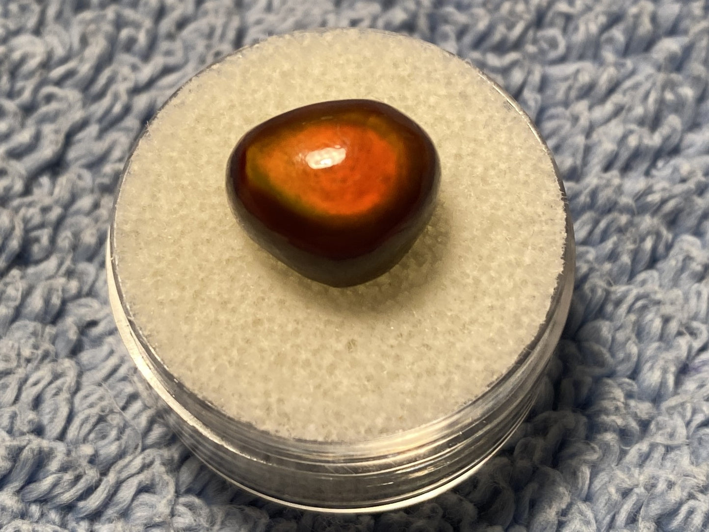 Round container with an orange colored Slaughter Mountain Fire Agate Gemstone on a textured blue surface