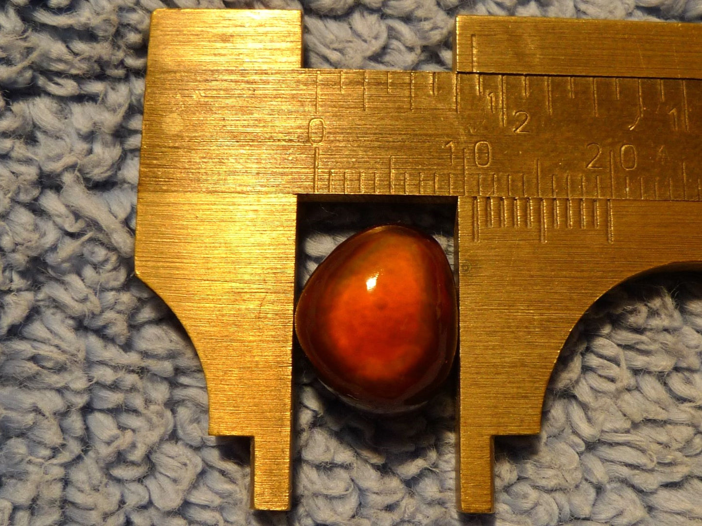 Gold caliper measuring a red-orange Slaughter Mountain Fire Agate Gemstone on a textured gray surface