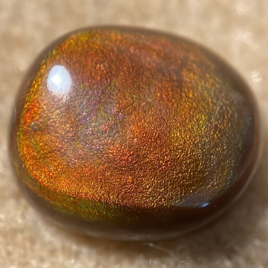 A polished, smooth oval gemstone with a rich iridescent color shift between coppery orange, gold, and deep brown, resting on a soft beige textured surface.