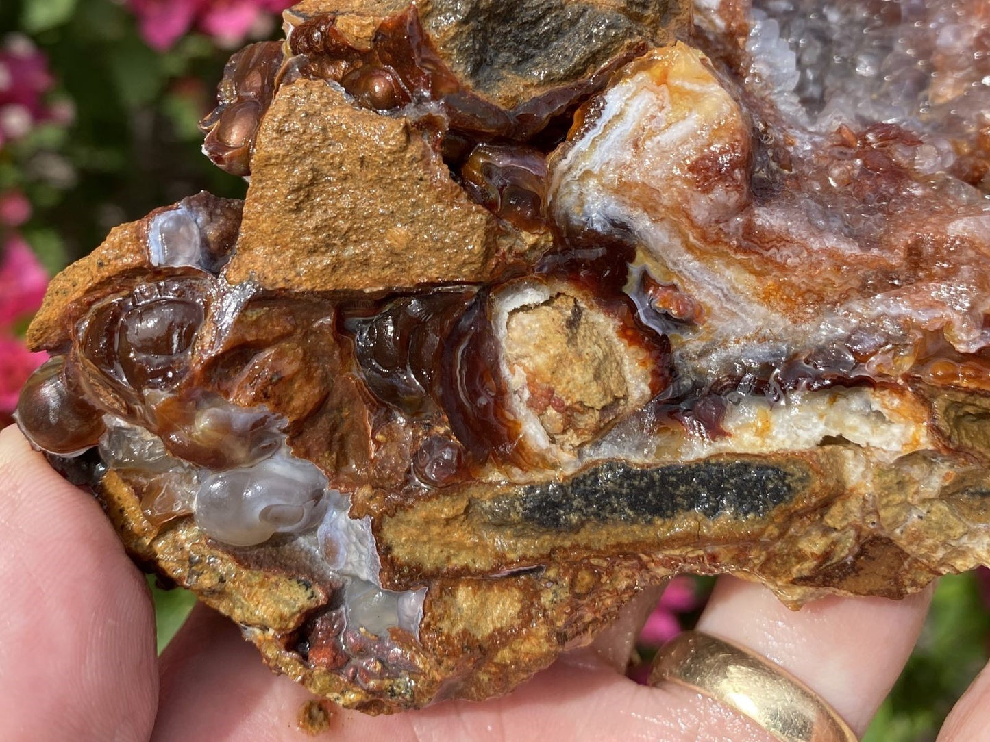 Close-up of a rock with crystals held between fingers, with blurred pink flowers in the background.