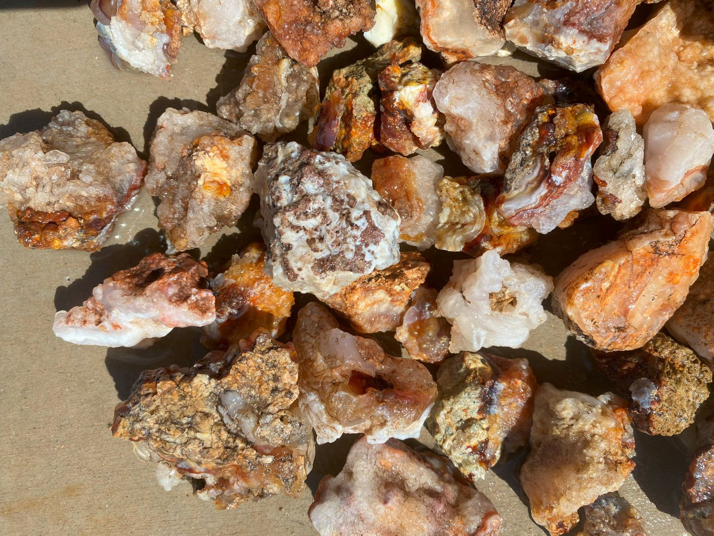A pile of rough, unpolished mineral stones and crystals in earthy tones of orange, brown, beige, and white, photographed closely on a tan surface.