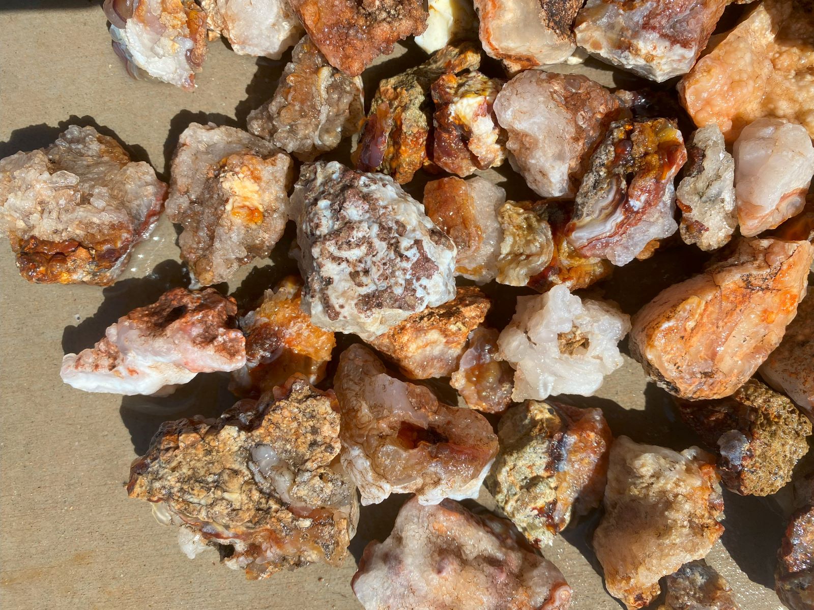 A pile of rough, unpolished mineral stones and crystals in earthy tones of orange, brown, beige, and white, photographed closely on a tan surface.
