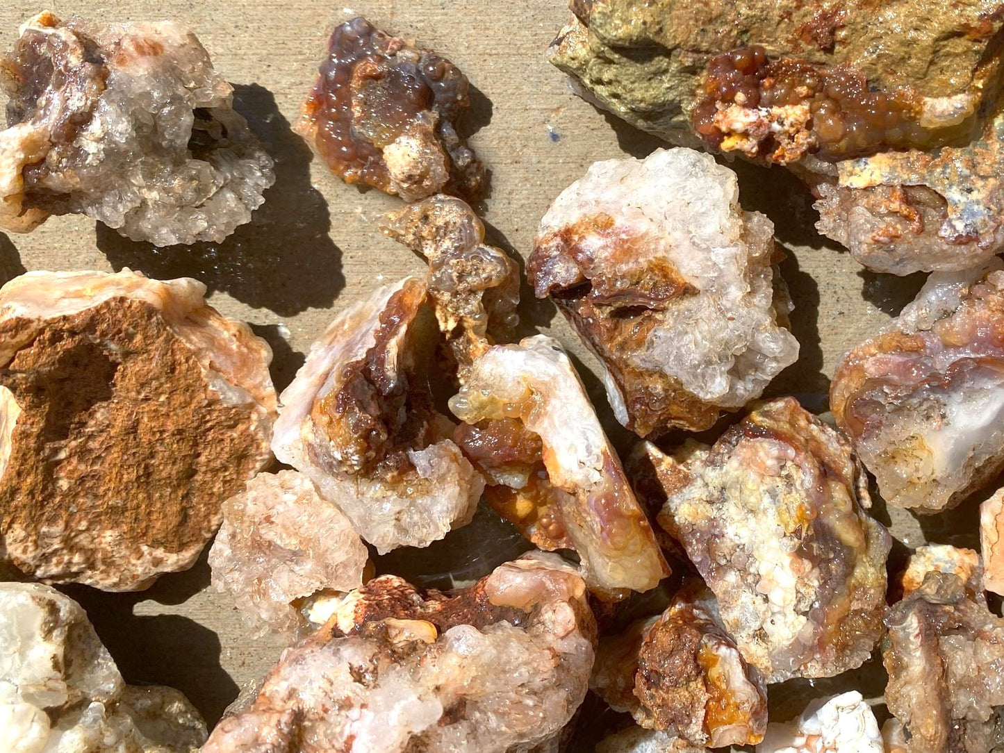 A close-up pile of rough mineral or stone chunks with a mix of translucent white, tan, brown, and orange hues on a sandy surface.