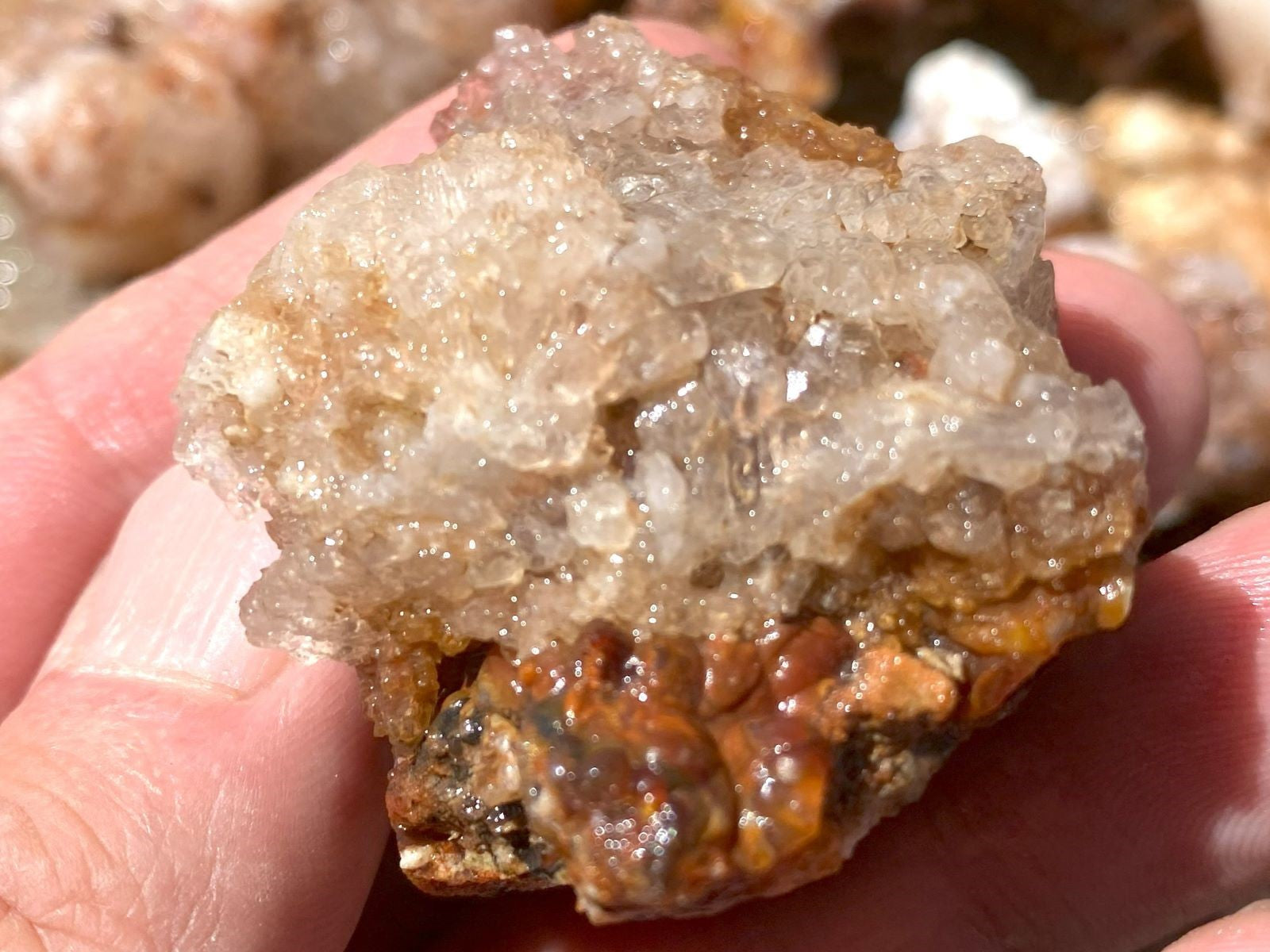 A close-up of a hand holding a raw crystal specimen with a translucent pale gray to off-white crystalline top and a brown and orange matrix base.