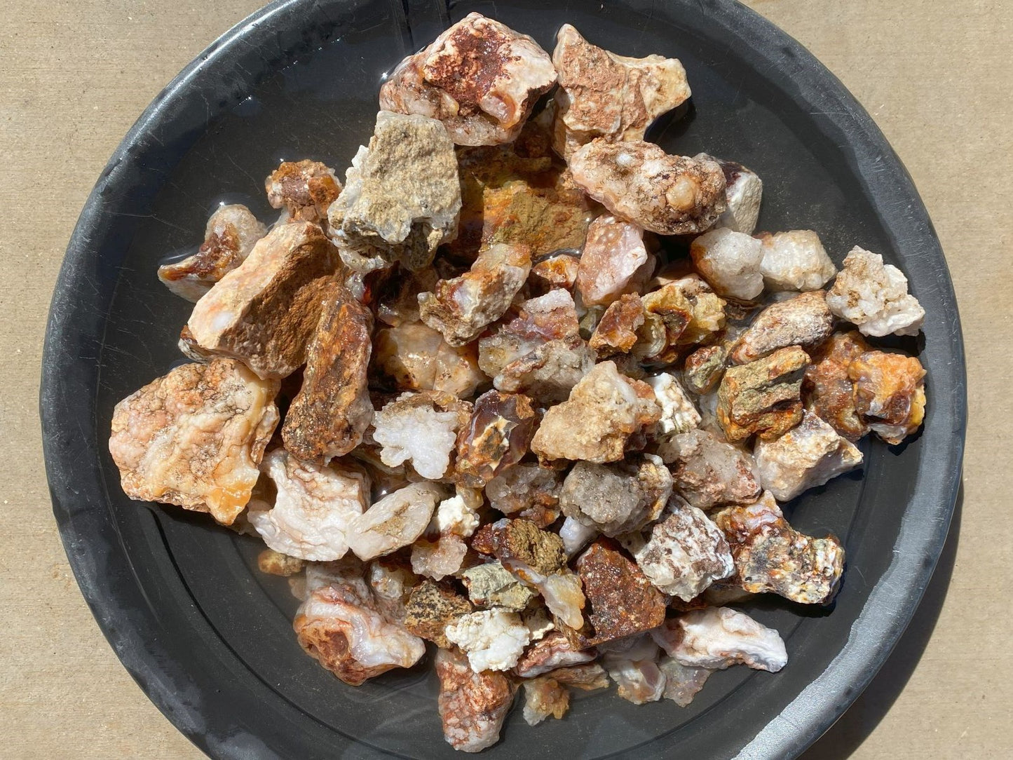 A pile of raw, rough mineral or stone chunks in earthy brown, beige, and white tones, displayed on a dark gray round plate against a tan background.