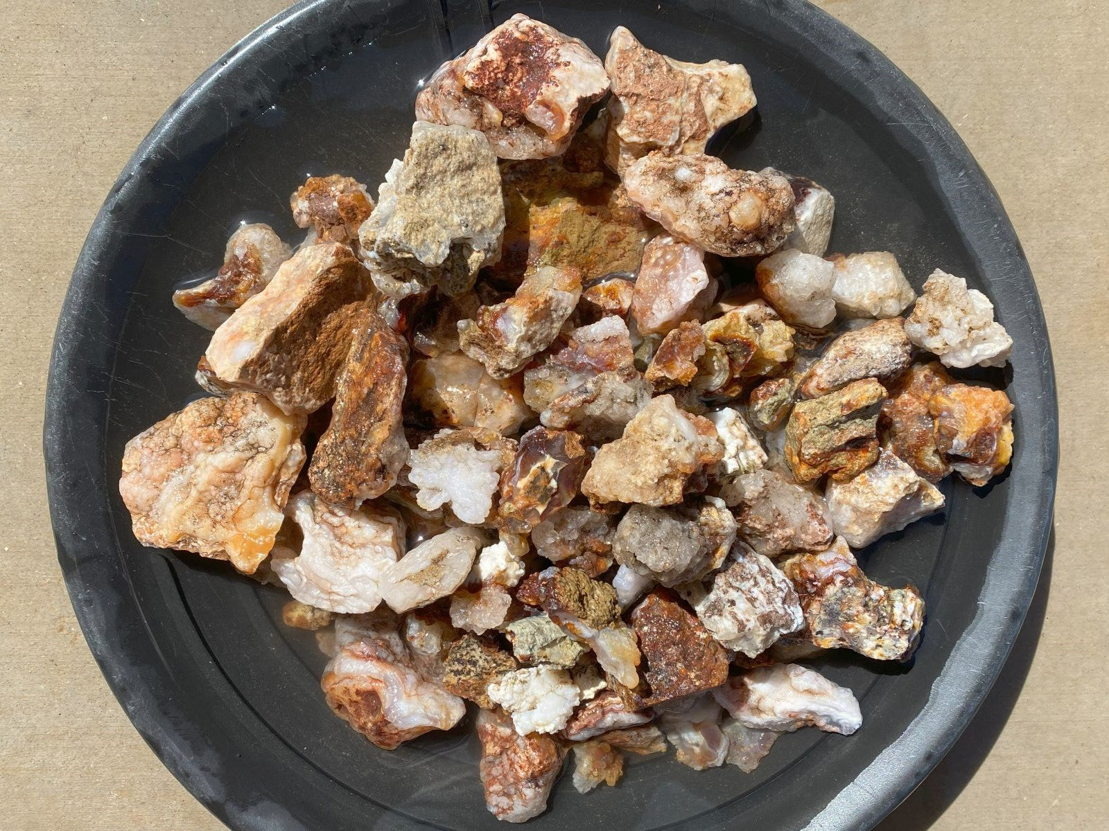 A pile of raw, rough mineral or stone chunks in earthy brown, beige, and white tones, displayed on a dark gray round plate against a tan background.