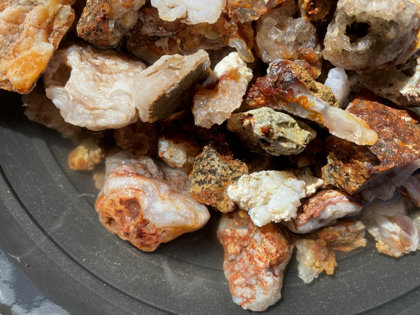 A pile of raw mineral specimens and crystals with varied textures, showing white agate, orange-brown quartz, and speckled stone pieces on a dark gray plate.