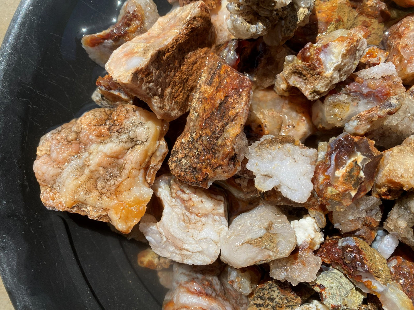 A close-up pile of raw mineral or stone chunks with rough, crystalline textures in beige, white, brown, and orange tones, resting in a black container.