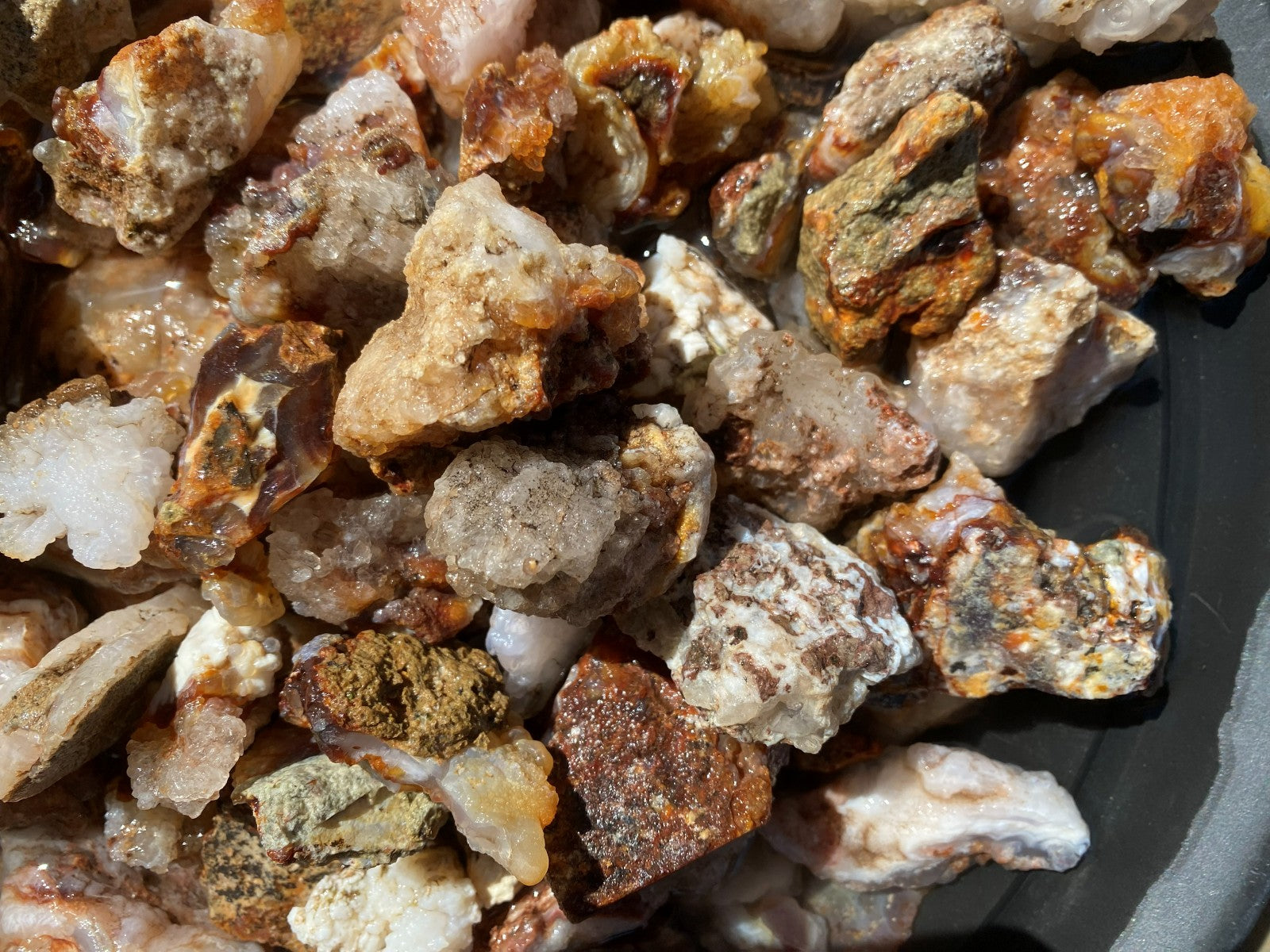 A close-up pile of rough, unpolished mineral or stone chunks in mixed earthy tones including tan, brown, gray, and white, filling the frame.