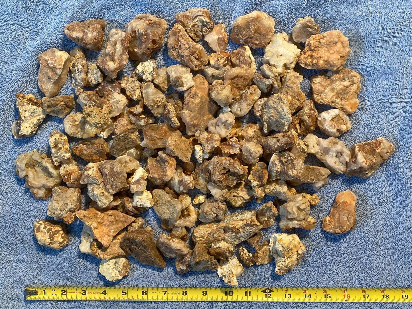 A pile of rough, raw mineral or stone chips in earthy brown, tan, and beige tones, spread out on a blue textured surface with a yellow tape measure visible at the bottom for scale.