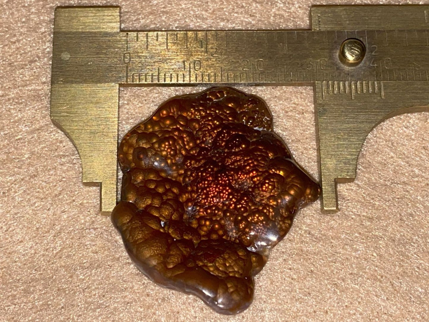 Fire Agate Gemstone being measured with a brass caliper on a beige surface
