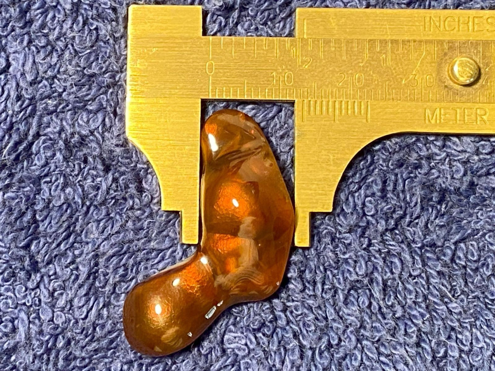 Orange colored fire agate stone being measured with a brass caliper on a textured blue surface