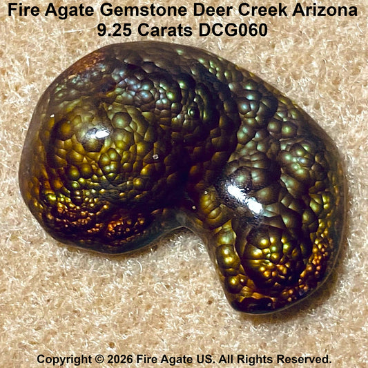 A polished Fire Agate gemstone featuring a pinfire bubble pattern of vibrant metallic gold, green, bronze and blue hues. The stone is displayed on a textured beige fabric background with text overlays identifying the source 'Deer Creek Arizona'.
