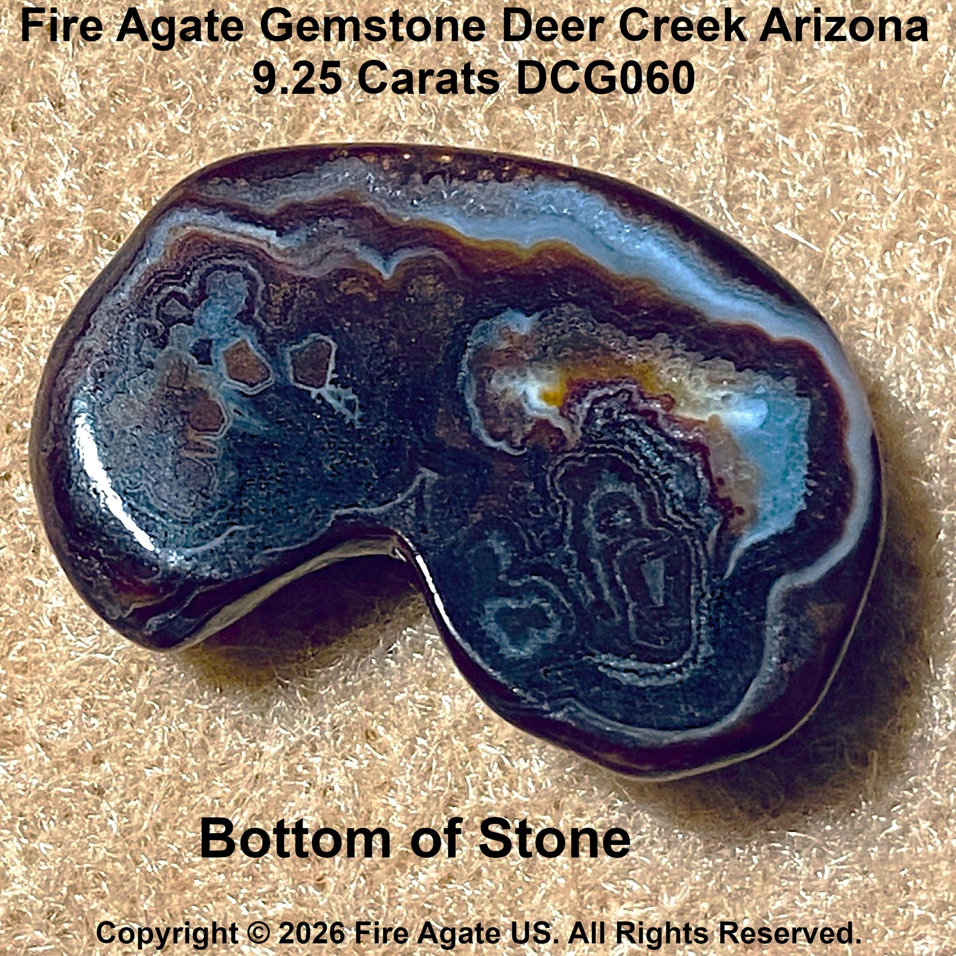 A polished bottom of a Fire Agate gemstone with a kidney-like shape, showing intricate banding in dark brown, black, light blue, and cream. The stone is photographed on a textured tan background with descriptive text overlays at the top and bottom.