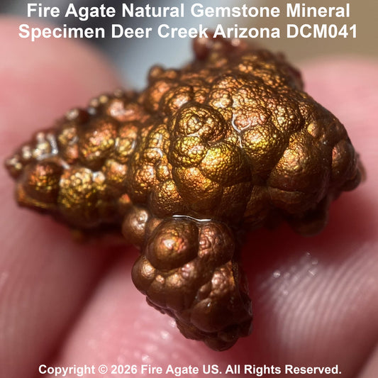 A close-up of a rough, bubbly fire agate gemstone specimen with metallic copper-gold tones, held against a blurred pink skin background. Text at the top reads 'Fire Agate Natural Gemstone Mineral Specimen Deer Creek Arizona DCM041' and a copyright notice appears at the bottom.