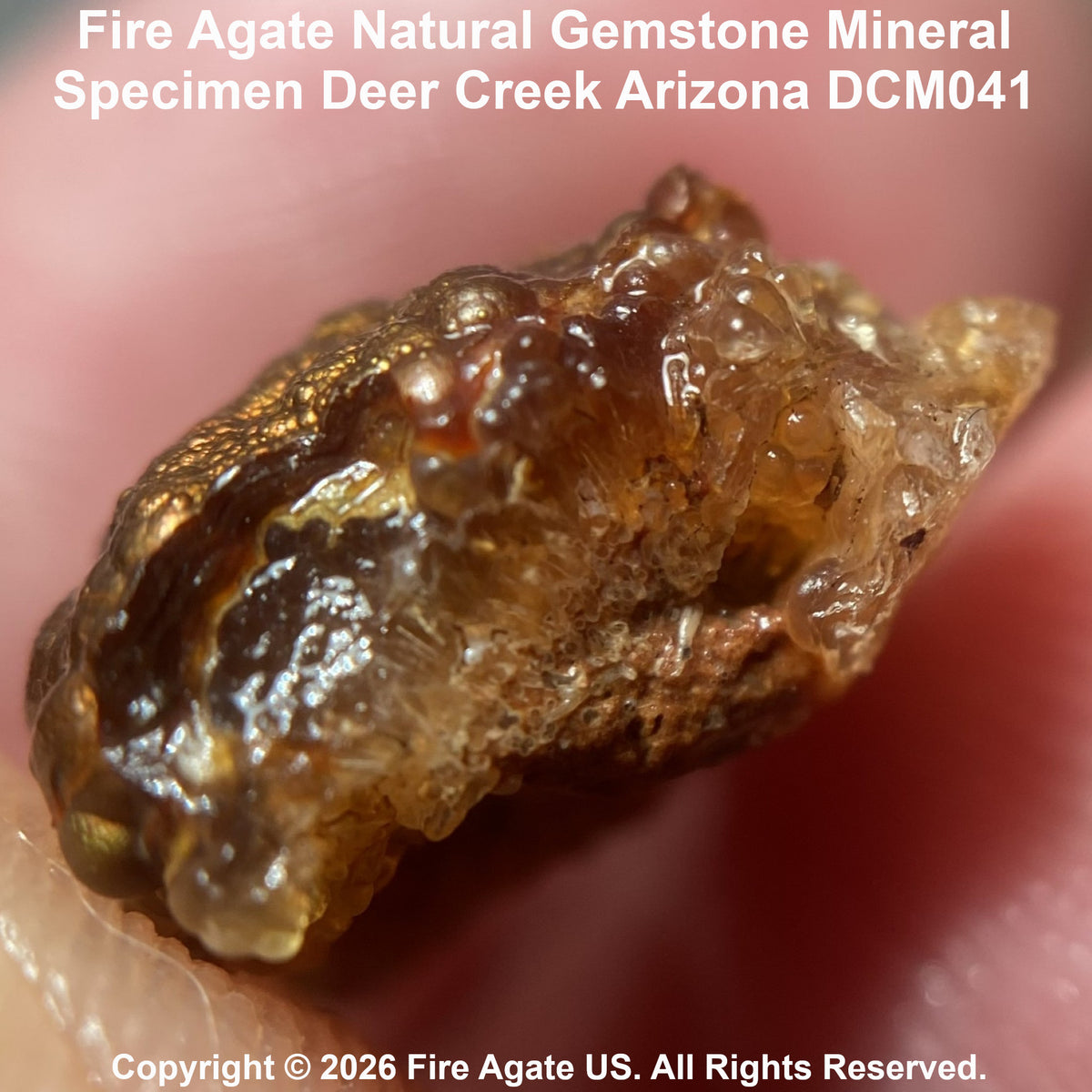 A close-up of a rough, natural fire agate gemstone mineral specimen held between fingers, with text at the top reading 'Fire Agate Natural Gemstone Mineral Specimen Deer Creek Arizona DCM041' and copyright text at the bottom.