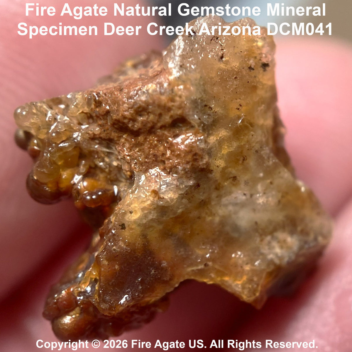 A close-up of a rough, translucent brown and amber fire agate gemstone specimen being held against a pink skin background, with text reading 'Fire Agate Natural Gemstone Mineral Specimen Deer Creek Arizona DCM041' and a copyright notice for Fire Agate US.