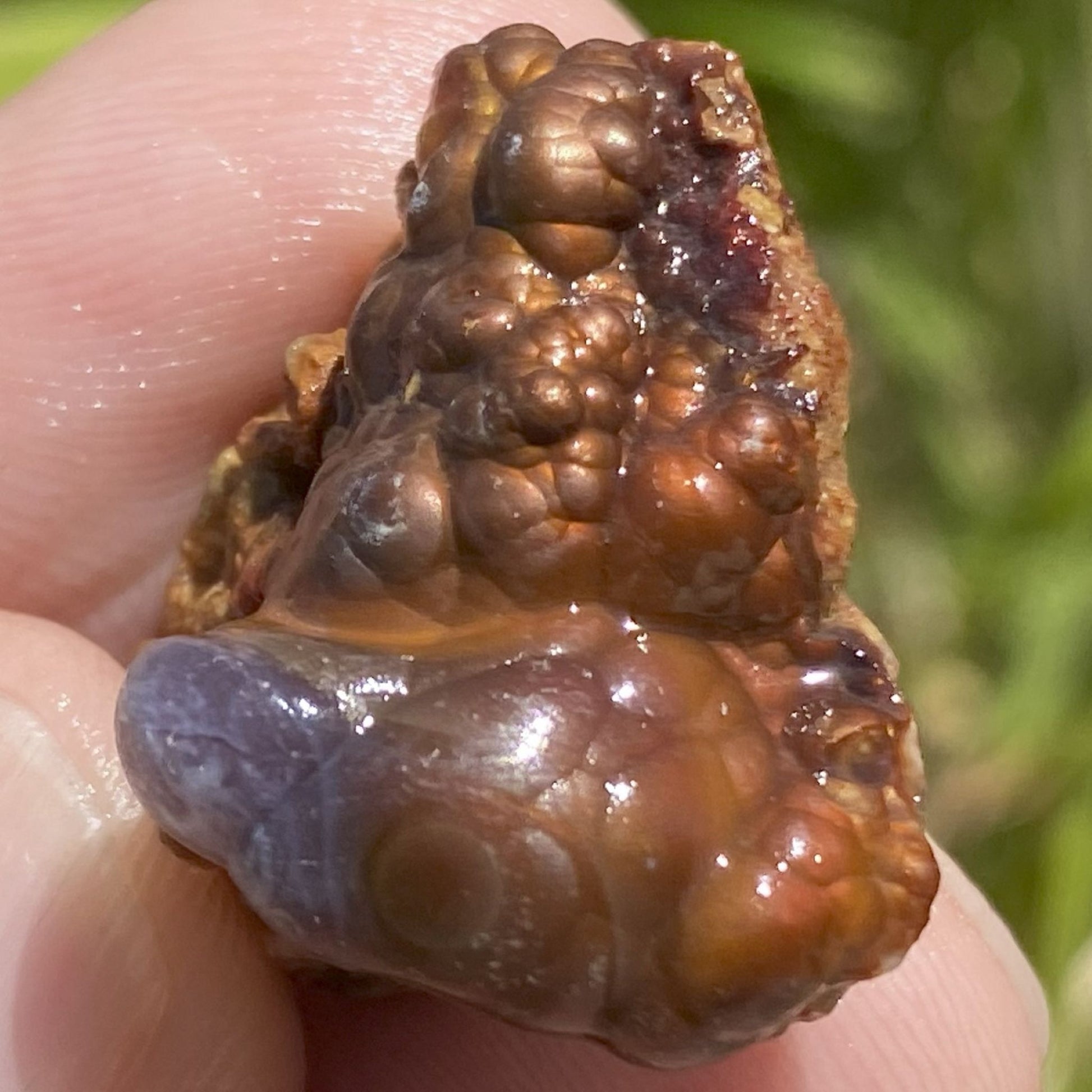 Close-up of a hand holding a Deer Creek fire agate rough stone against a blurred natural background.