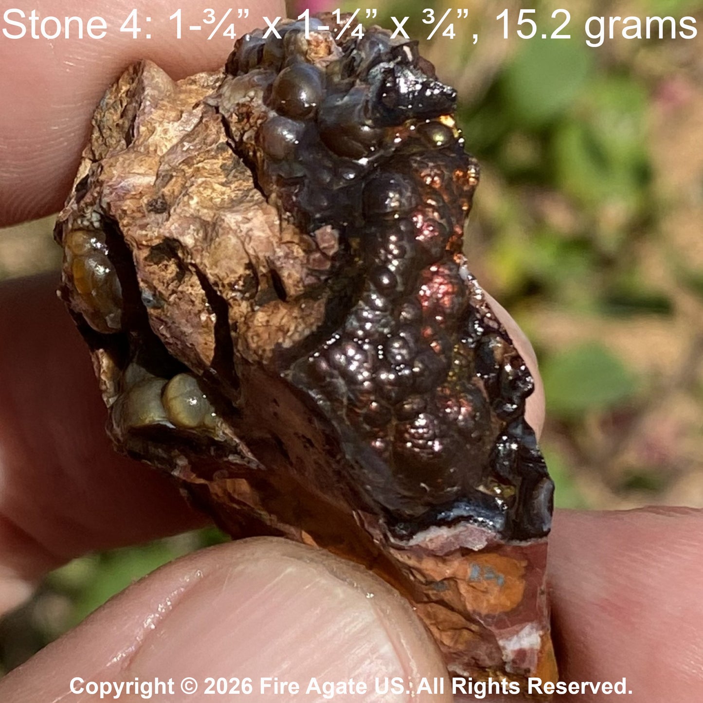 A hand holding a rough fire agate stone specimen with brown, black, and coppery metallic looking inclusions on a tan stone matrix, outdoors with blurred greenery in the background. Text at top reads 'Stone 4: 1-3/4 x 1-1/4 x 3/4, 15.2 grams'