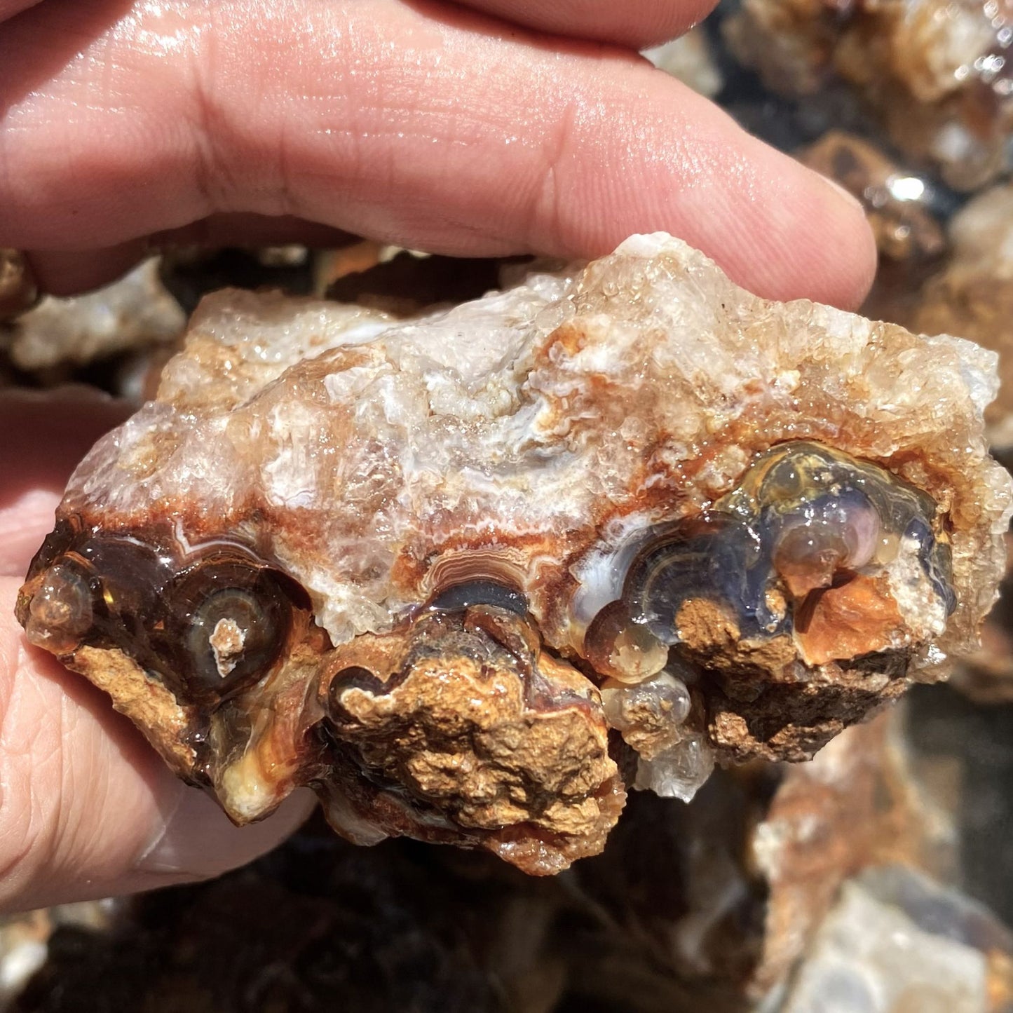 A hand holding a piece of fire agate rough or crystal specimen with layered translucent brown, amber, and smoky gemstone formations against a blurred outdoor rocky background.