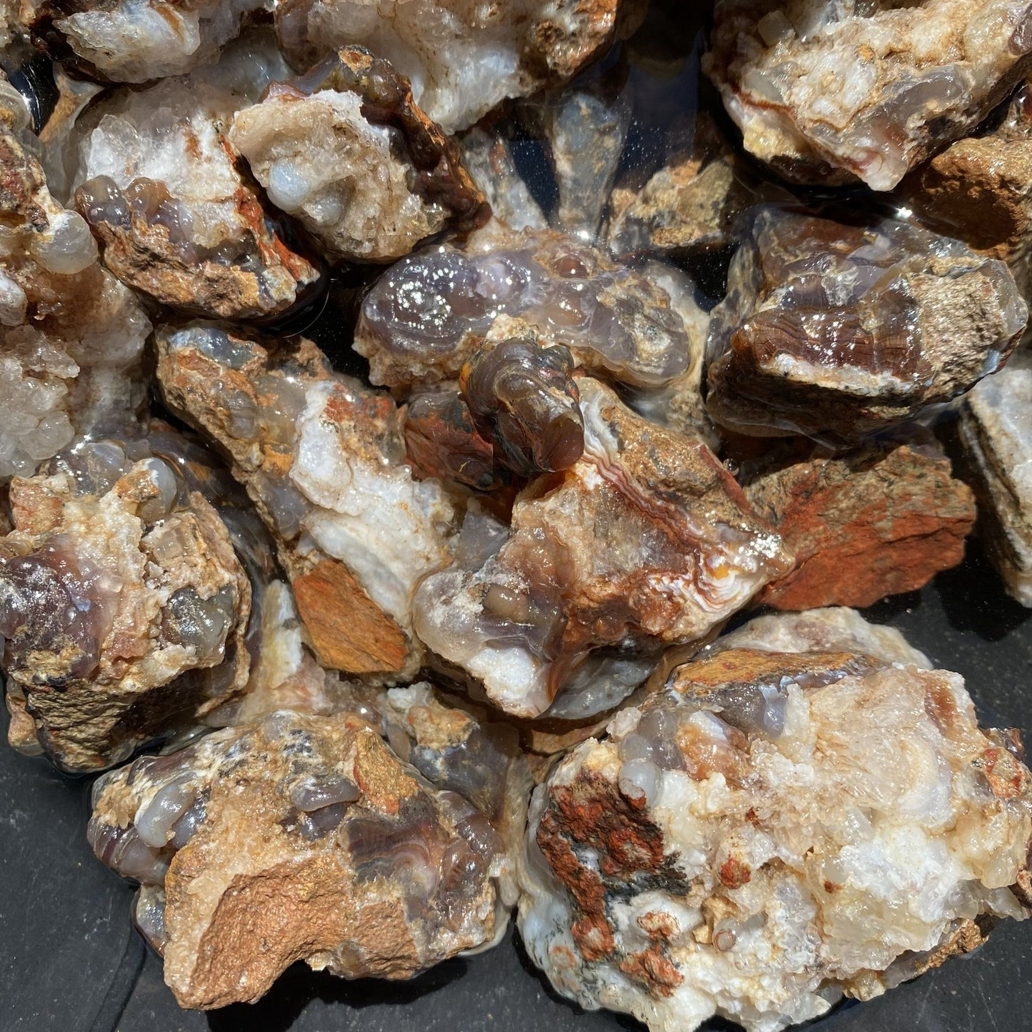 A pile of rough agate and quartz crystal clusters with natural matrix, showing translucent bands of gray, brown, orange, and white.