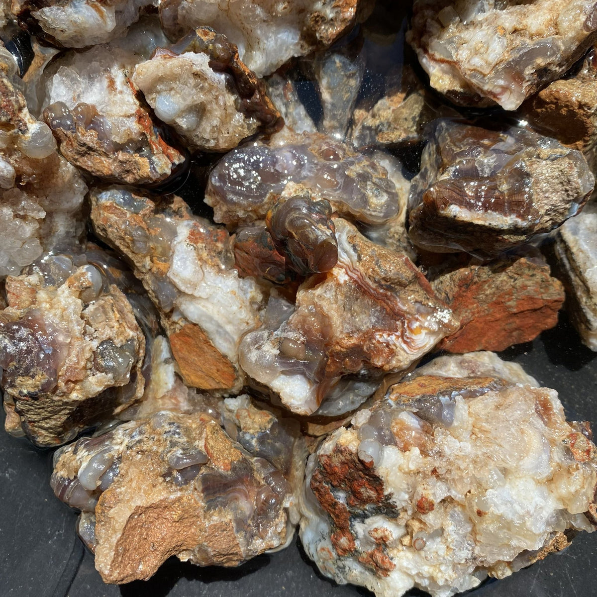 A pile of rough agate and quartz crystal clusters with natural matrix, showing translucent bands of gray, brown, orange, and white.