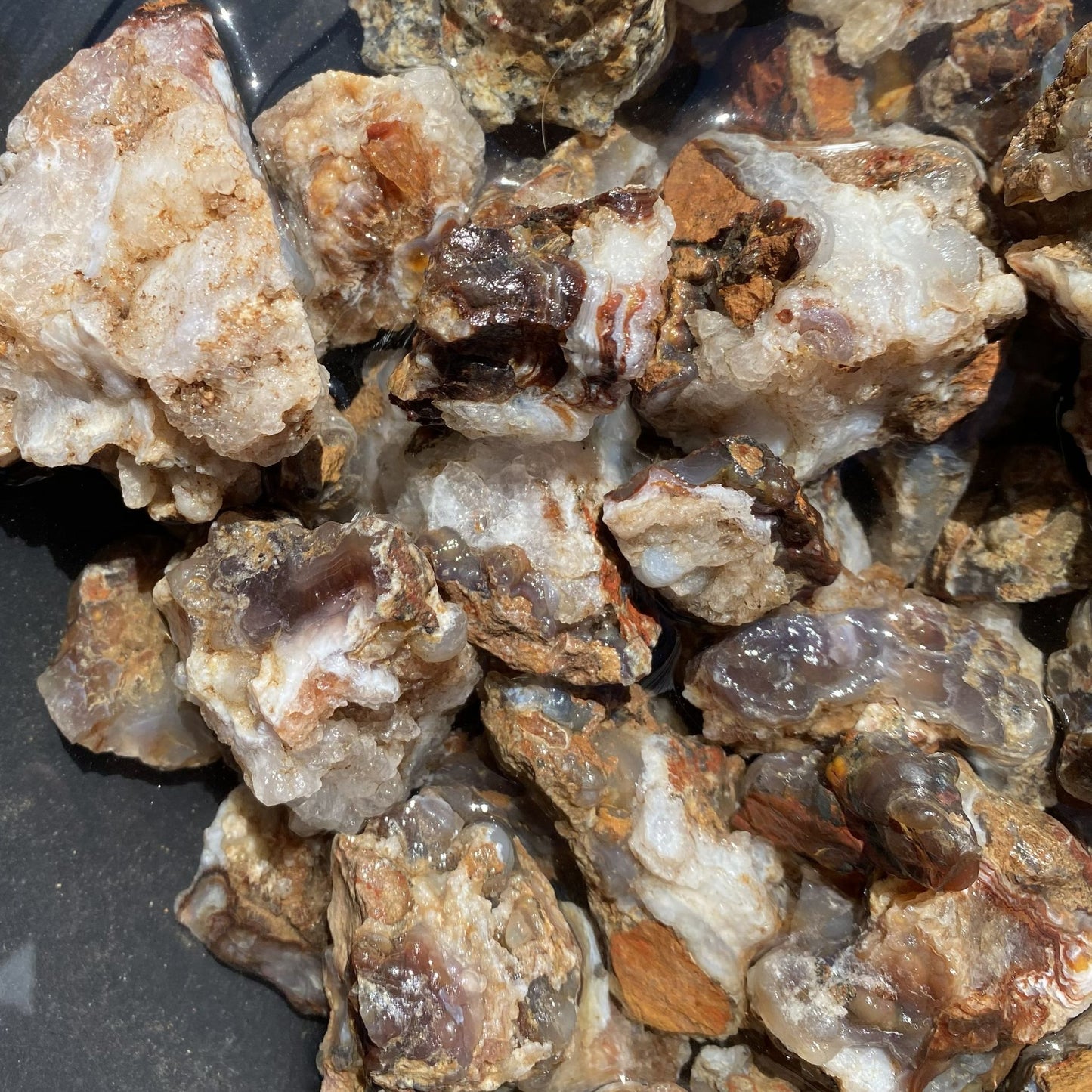 A close-up pile of rough fire agate and quartz stone chunks with natural matrix, showing translucent white, gray, brown, and amber tones on a dark surface.
