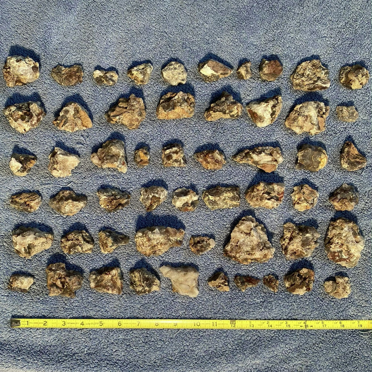 A group of irregular, rough, tan and brown fire agate pieces arranged in rows on a blue textured surface, with a yellow measuring tape along the bottom edge for scale.