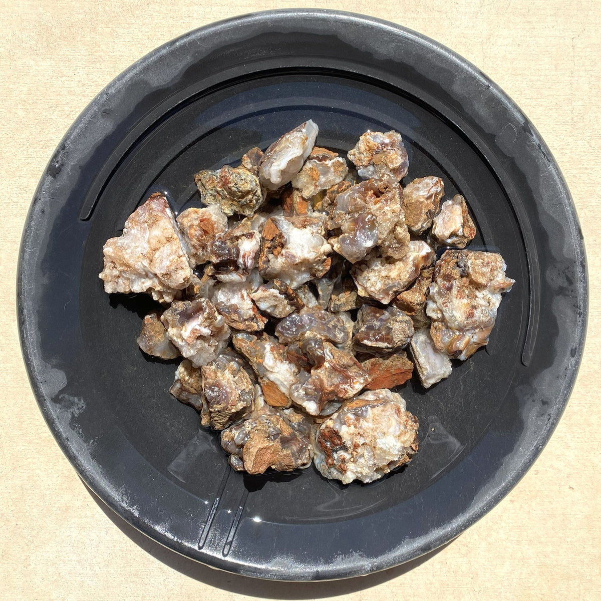 A pile of irregularly shaped natural stone fire agates with earthy brown, beige, and gray tones, displayed inside a black plastic bowl on a light tan textured background.