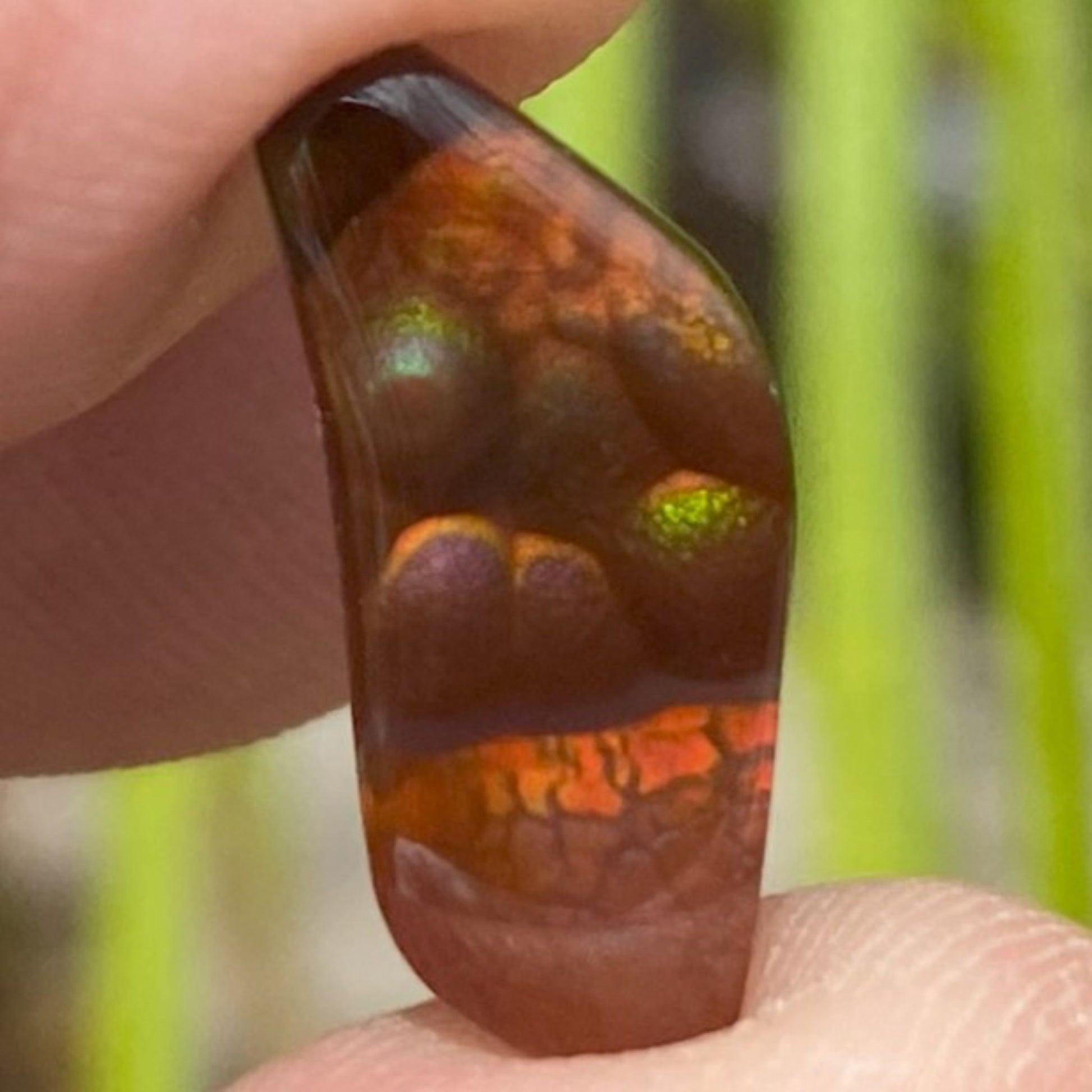 A close-up of a polished brown fire agate gemstone held between fingers, showing iridescent green and orange play-of-color against a blurred green outdoor background.
