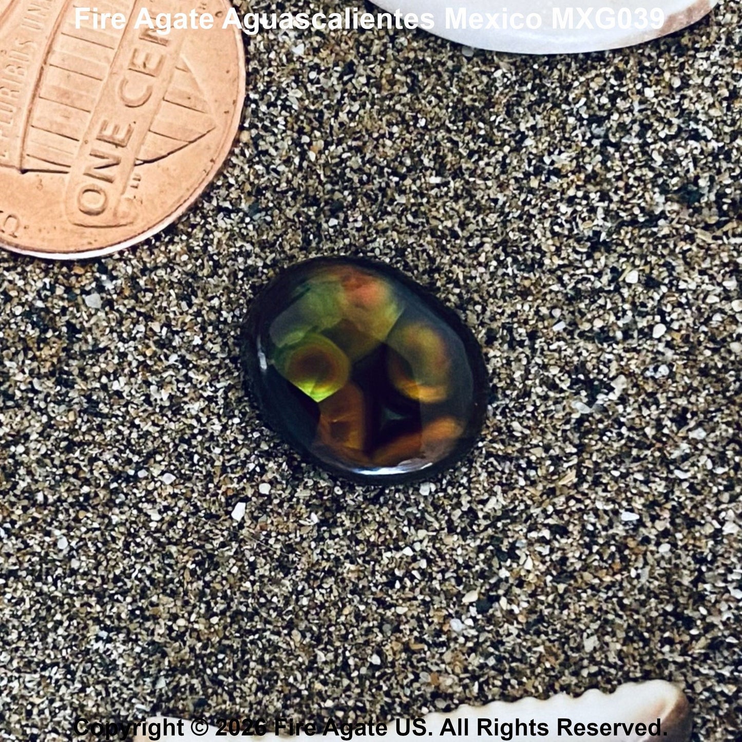 A polished oval Fire Agate gemstone with a dark base and vibrant green, yellow, and orange flashes, resting on coarse sand next to a US penny for scale.