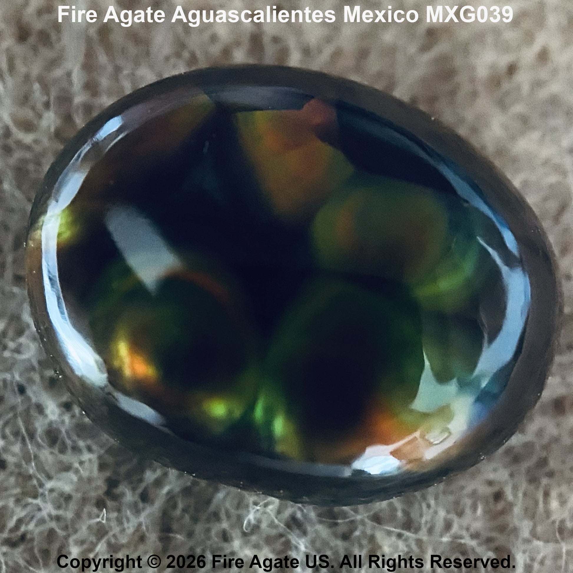 A polished Fire Agate gemstone cabochon showing a mix of dark green, black, and fiery orange-yellow flashes, resting on a textured beige surface. Text overlays identify the stone as 'Fire Agate Aguascalientes Mexico MXG039' with a copyright notice for 'Fire Agate US'.