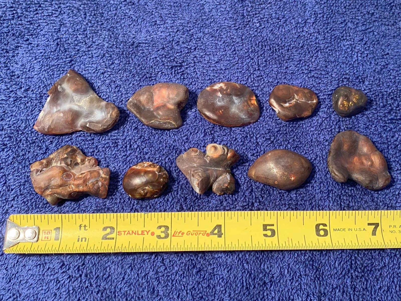 Collection of 10 fire agate gemstone preforms on a blue surface with a measuring tape for scale.