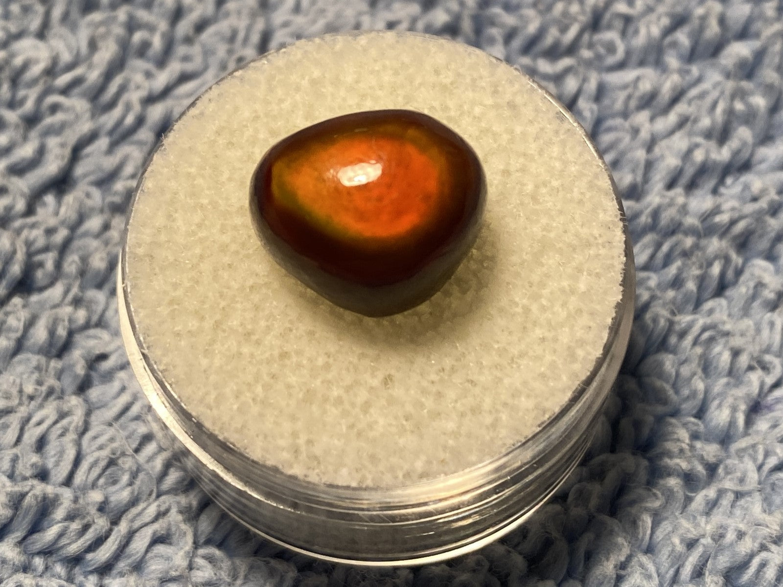 Round container with an orange colored Slaughter Mountain Fire Agate Gemstone on a textured blue surface