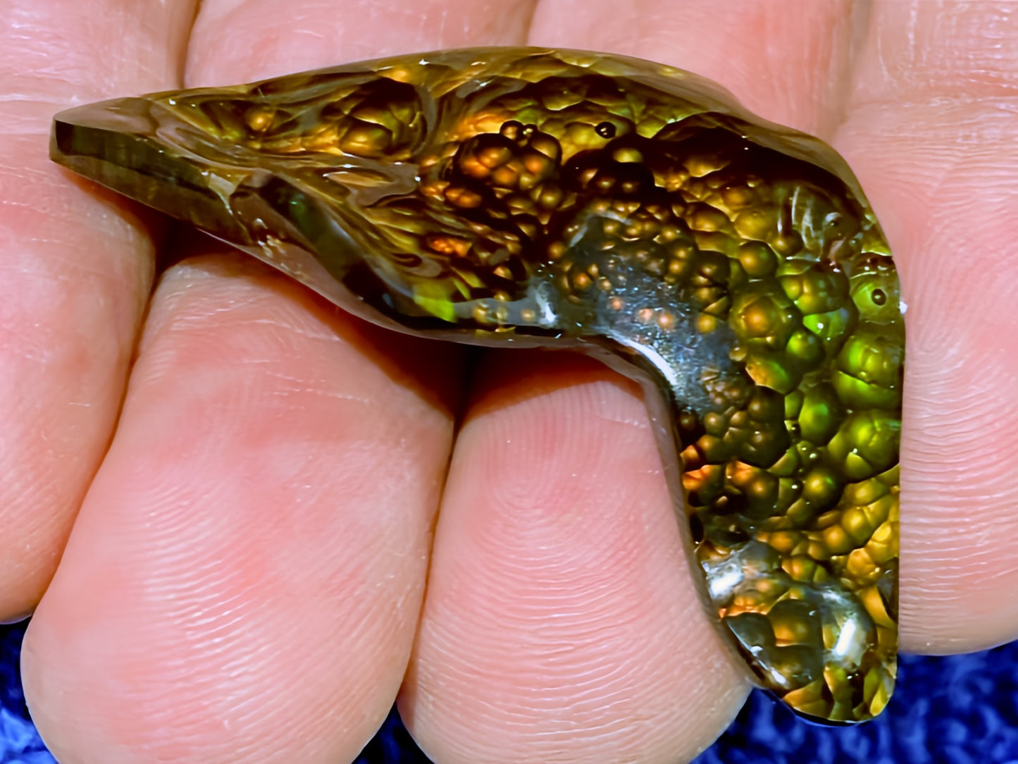 Close-up of a hand holding a green and orange colored loose fire agate gemstone with intricate fire patterns.