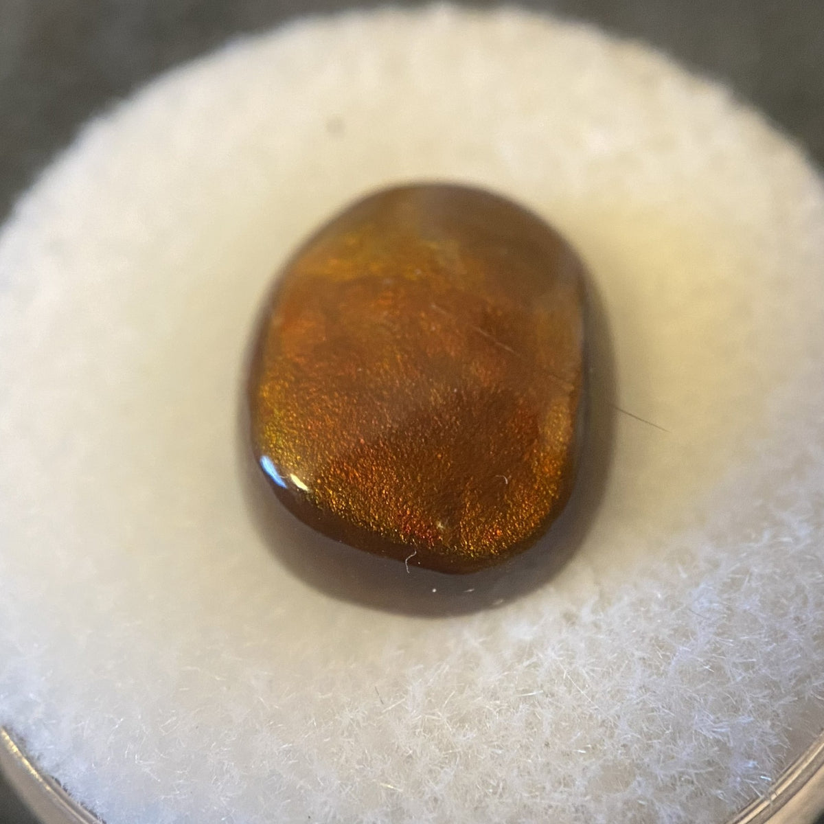 A polished, teardrop-shaped gemstone with a metallic coppery-brown sheen and iridescent shimmer, resting on a white textured foam surface.