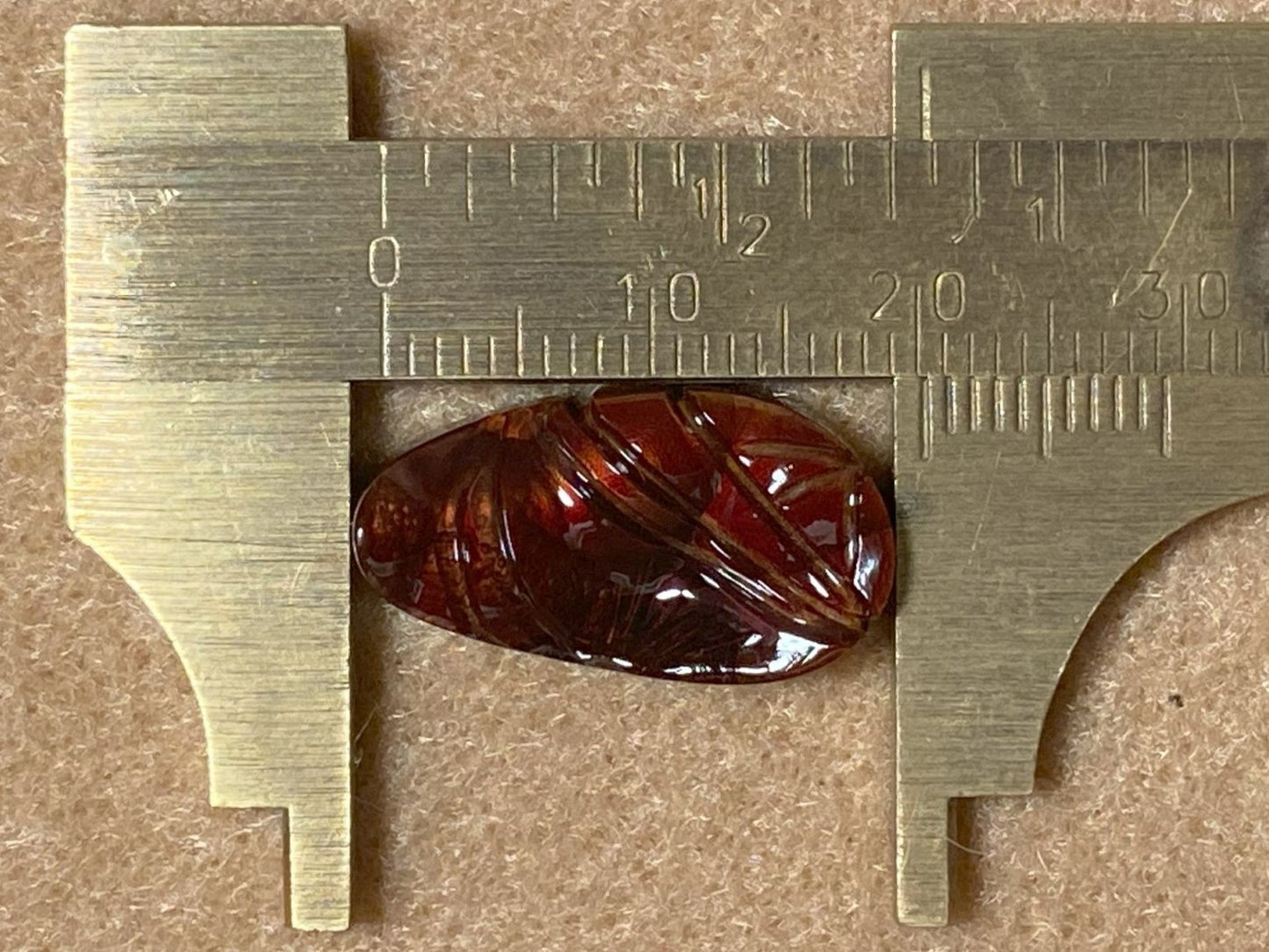 Burgundy colored fire agate gemstone being measured with a brass caliper on a brown background
