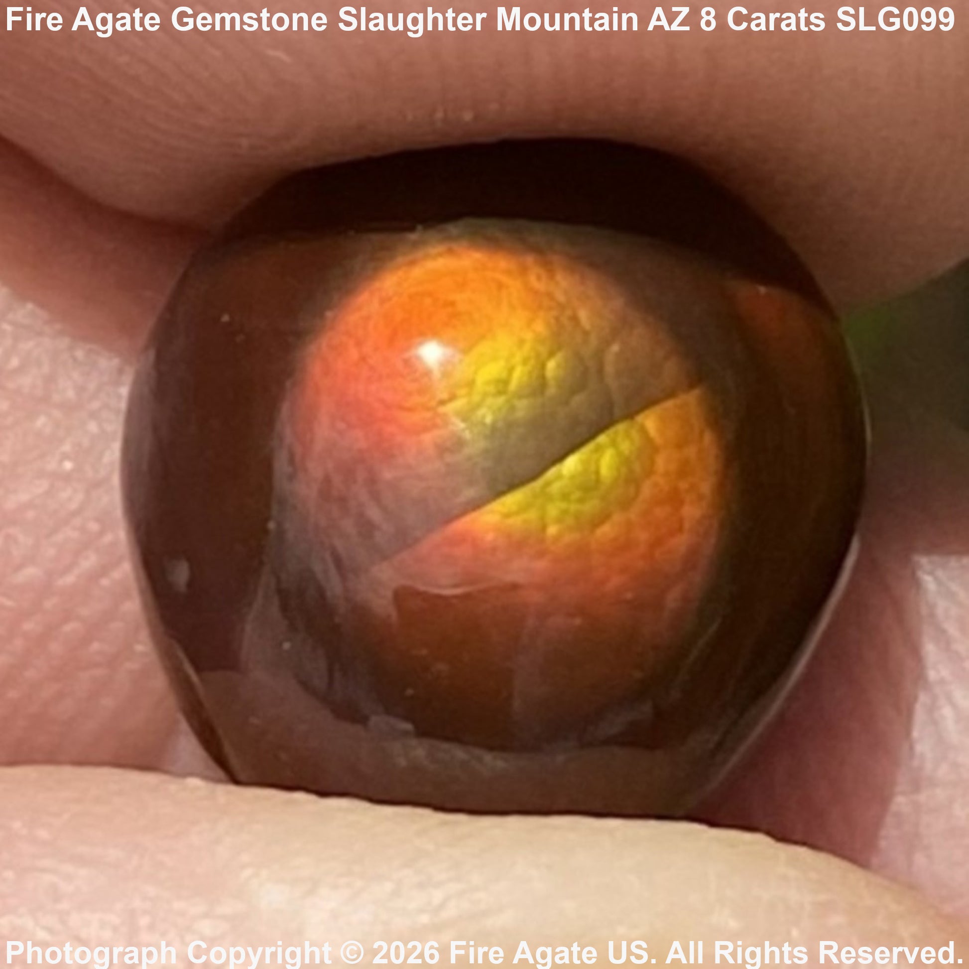 A close-up of a polished fire agate gemstone held between fingers, showing a strong rainbow iridescent play-of-color with orange, yellow, and green reflections on a dark brown base. Text overlays identify the stone as 'Fire Agate Gemstone Slaughter Mountain AZ 8 Carats SLG099' and credit 'Fire Agate US'.