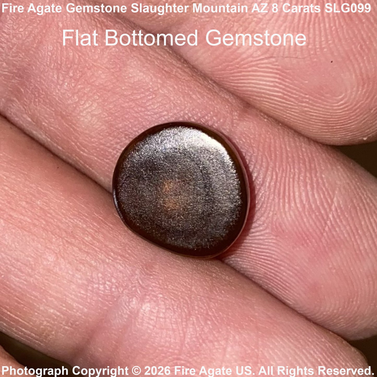 A flat, round cabochon gemstone with a metallic dark brown and silver-gray finish, held on a person's fingers to show its size. Text overlays identify the gemstone as Fire Agate and specify 'Flat Bottomed Gemstone'.