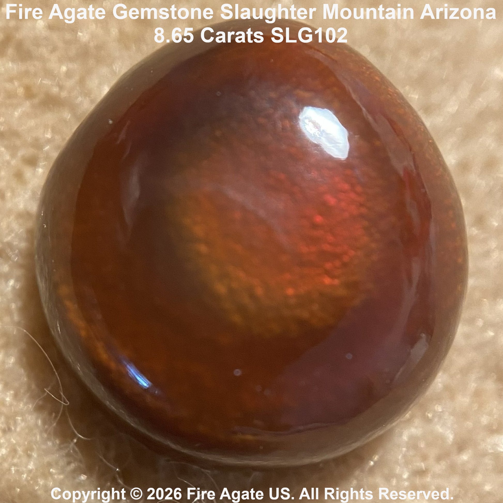 A polished fire agate gemstone with a rounded, dome-like shape, showcasing a warm brown body with iridescent flashes. The stone is photographed close-up on a textured beige surface, with text overlays at the top and bottom identifying it as 'Fire Agate Gemstone Slaughter Mountain Arizona'.