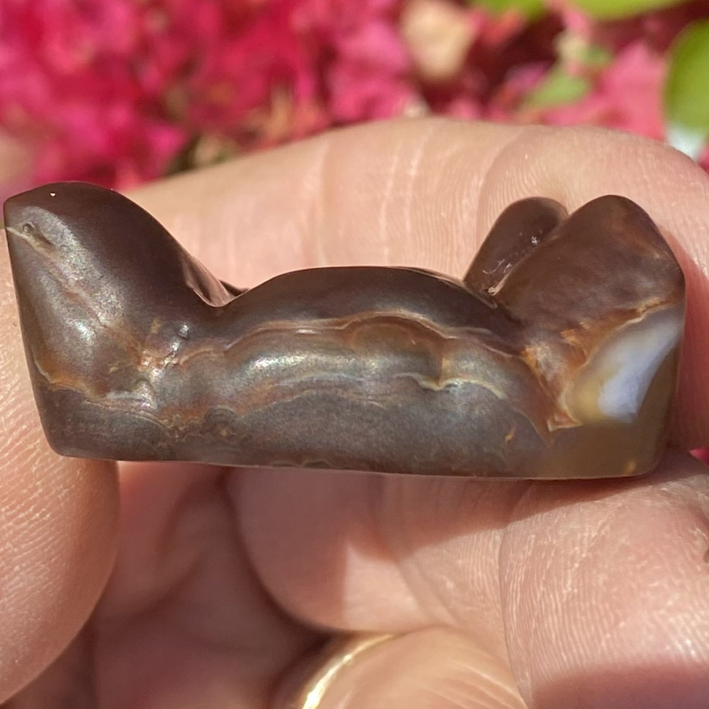 Slaughter Mountain preform fire agate gemstone with wavy texture held between fingers against a blurred pink floral background