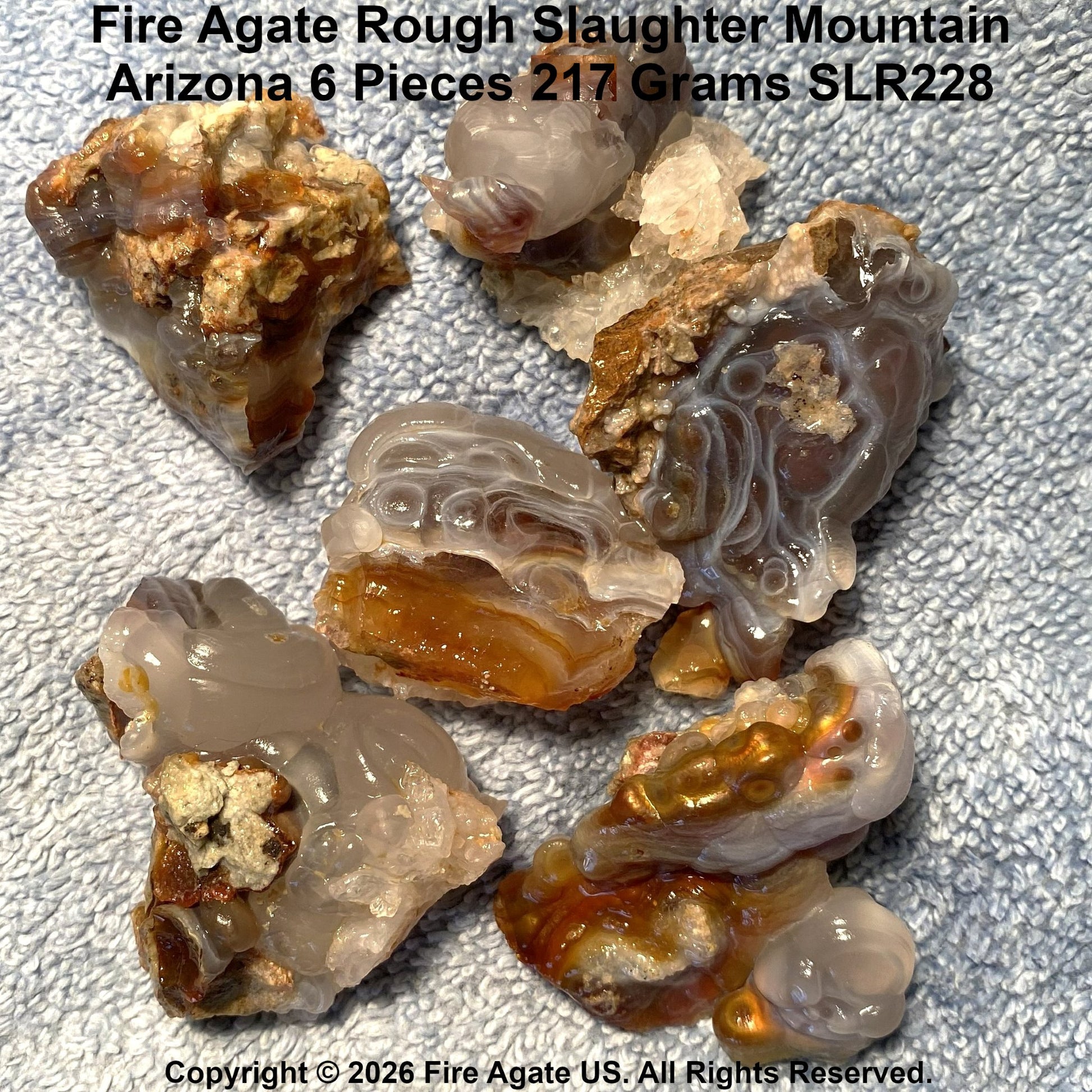 Six pieces of rough fire agate from Slaughter Mountain, Arizona, displayed on a textured fabric background. The agate pieces show natural banding in translucent shades of gray, beige, and amber brown, with some pieces showing metallic coppery mineral inclusions.
